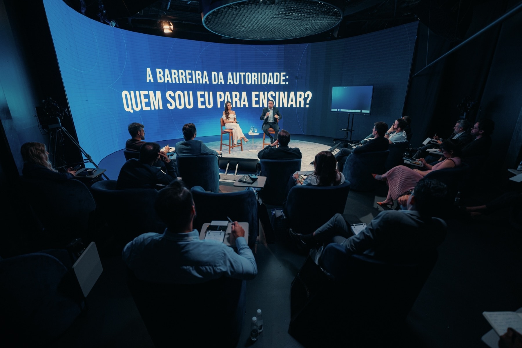 Palco do evento com o telão curvo aceso e a plateia em poltronas em volta, durante uma conversa entre dois palestrantes